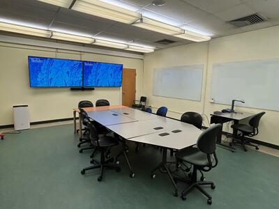 Classroom with two monitors, large table w/ 11 chairs, instructor table